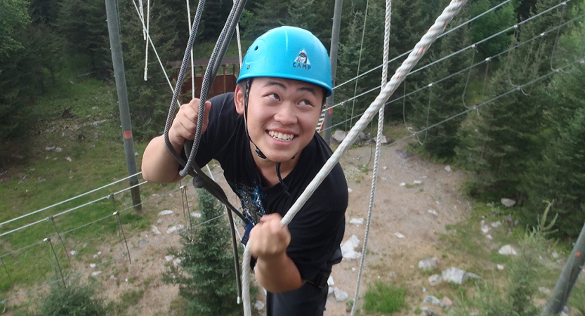 A person wearing safety gear and secured by ropes smiles as they navigate an obstacle of a high ropes course. 